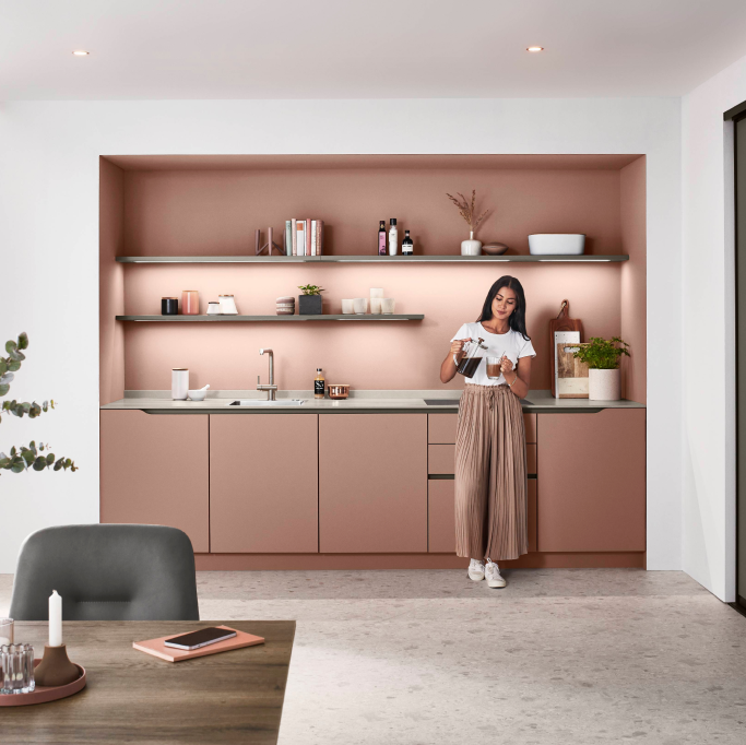 Eine Frau steht in einer pinken Küche und lächelt, umgeben von modernen Küchengeräten